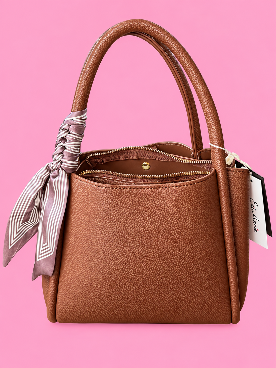 A brown pebbled leather handbag with double top handles, a zippered top, and a striped satin scarf tied to one handle, shown against a solid pink background.