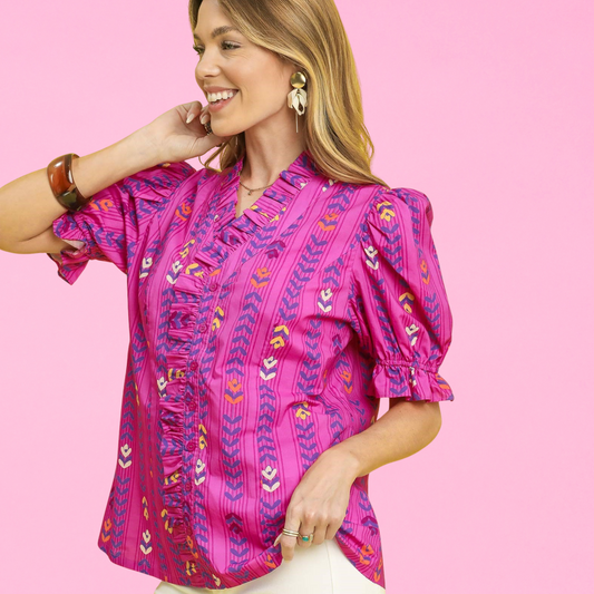 Woman smiling in a magenta puff-sleeve blouse with ruffled neckline and button front, styled with white pants and statement earrings against a yellow background.