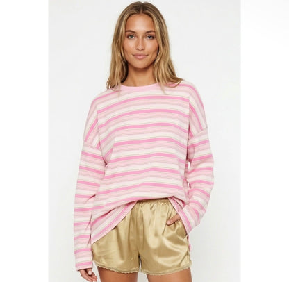 A woman wearing a pink and white striped long-sleeve crewneck sweatshirt and shiny gold satin shorts, standing against a plain light gray studio background.