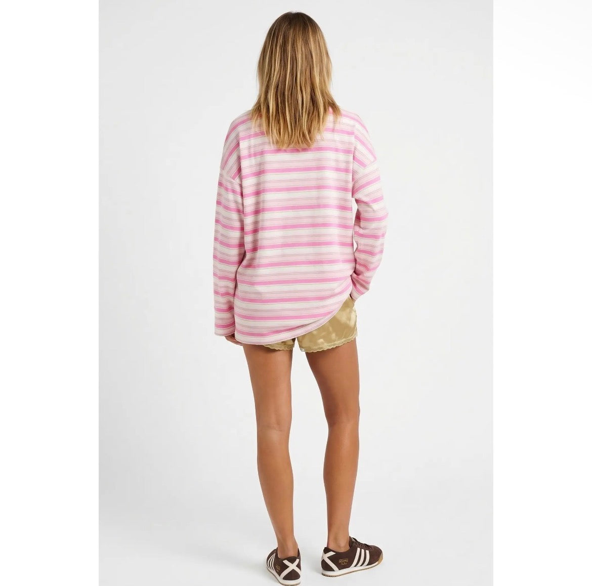 A woman viewed from behind wearing a pink and white striped long-sleeve t-shirt, gold patterned shorts, and brown sneakers against a light gray studio background.