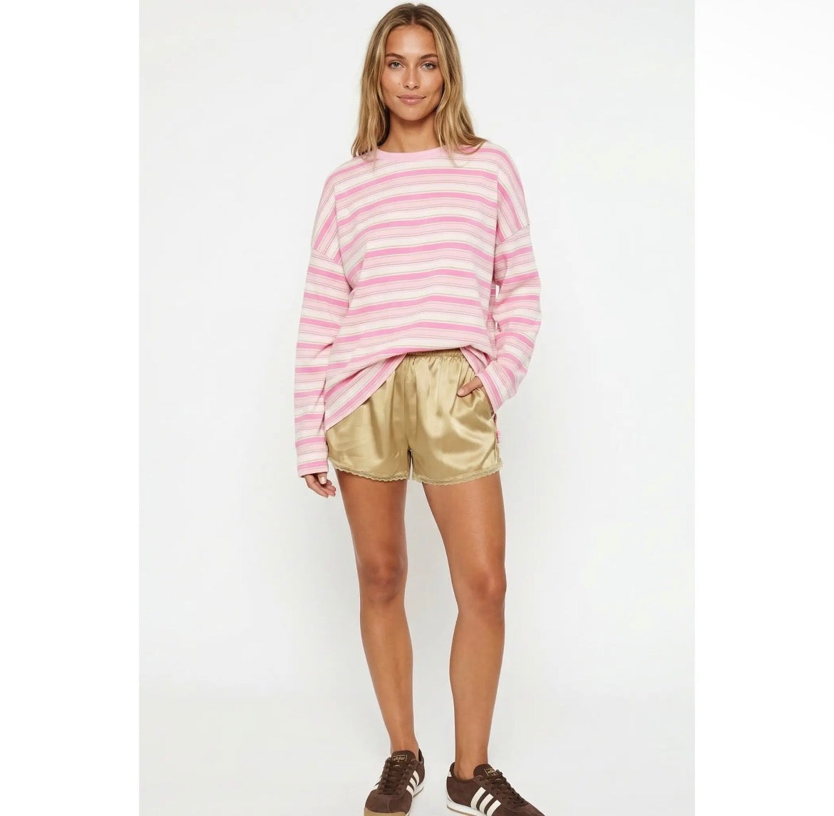 A woman modeling a pink and white horizontally striped long-sleeve knit top with metallic gold satin shorts and brown sneakers, standing against a plain white studio background.