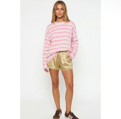 A woman modeling a pink and white horizontally striped long-sleeve knit top with metallic gold satin shorts and brown sneakers, standing against a plain white studio background.