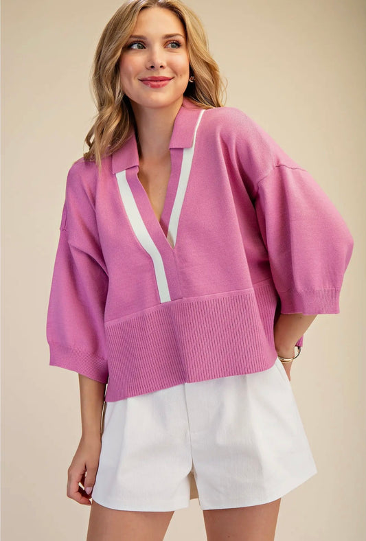 A woman wearing a pink knit collared sweater with white contrast trim and white shorts, photographed against a plain beige studio background.