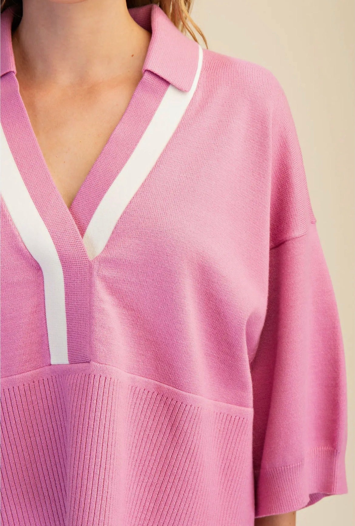 A close-up of a pink knit polo-style top with a V-neck, featuring white contrast panels running down the front and along the collar, worn by a model.