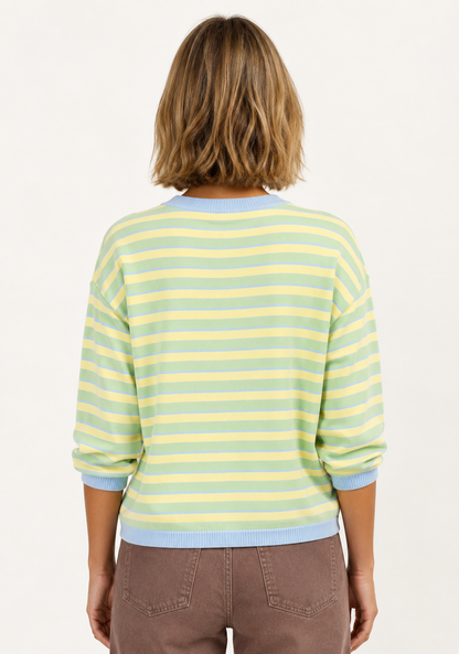 A woman viewed from behind wearing a pastel striped knit sweater with light blue and pale yellow horizontal stripes, paired with brown pants, against a plain white background.