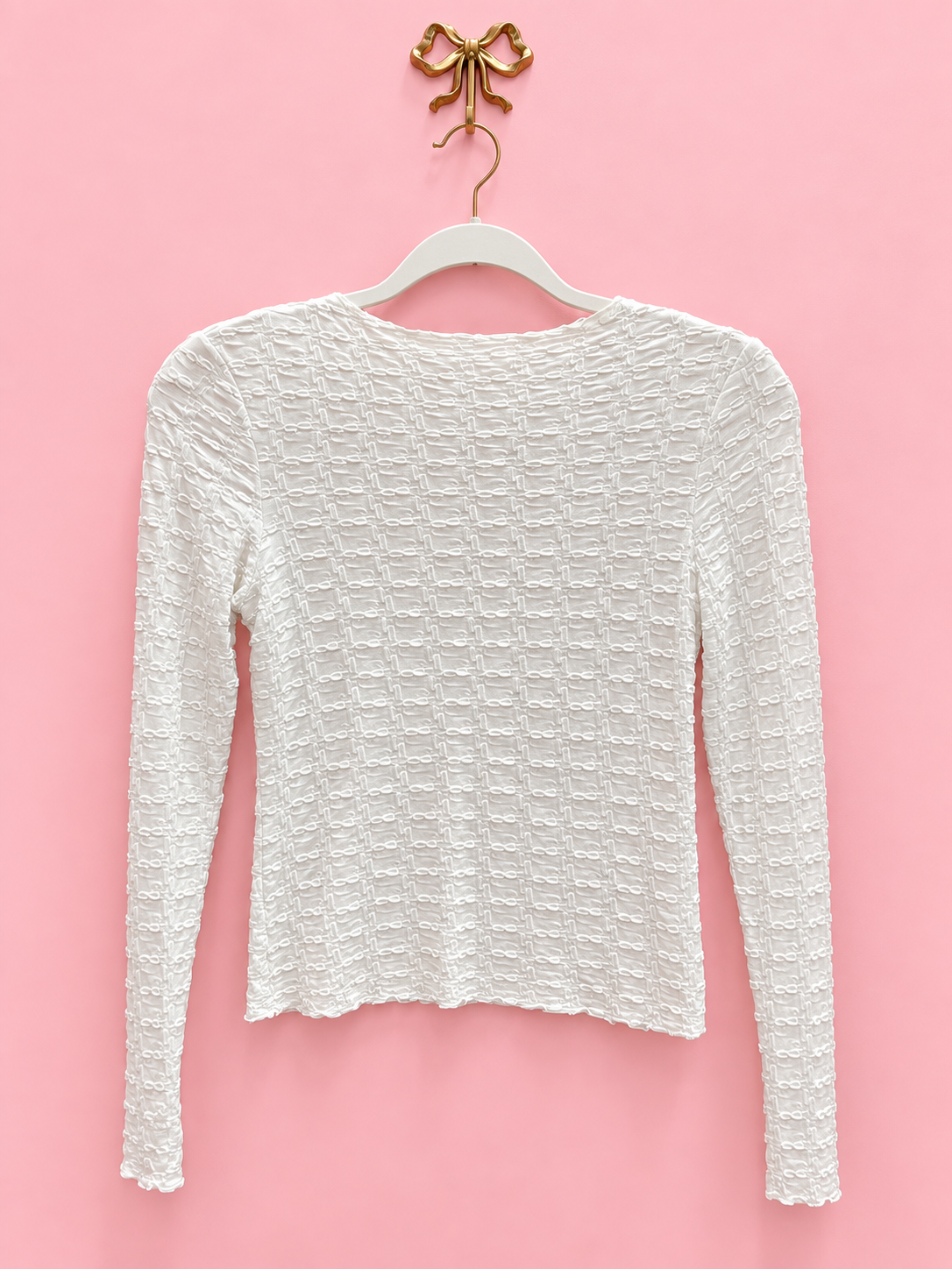 A white textured long-sleeve top hanging on a white hanger with a gold bow-shaped hook, set against a solid pink background.