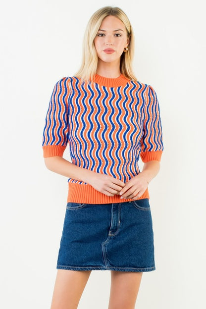 A woman modeling a short-sleeve knit sweater with a wavy blue, orange, and white pattern and solid orange ribbed trim, paired with a blue denim mini skirt against a plain white background.