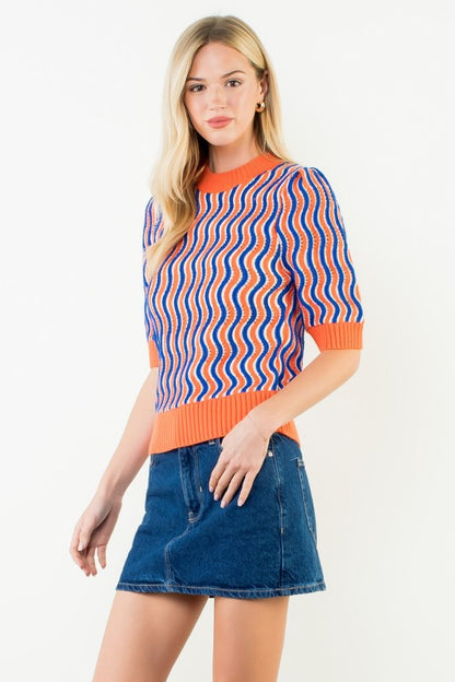A woman modeling a short-sleeve knit sweater with vertical wavy stripes in orange, blue, and white, paired with a blue denim mini skirt, against a plain white studio background.