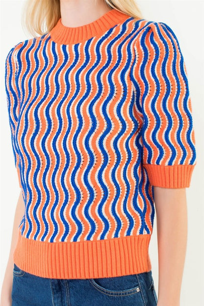 A close-up of a model wearing a short-sleeve knit sweater with a vibrant wavy stripe pattern in orange and blue, paired with blue jeans.