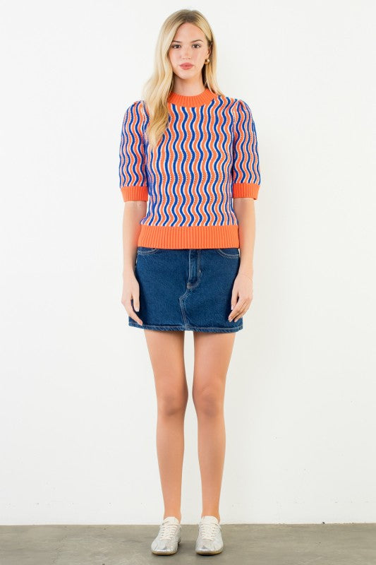 A woman modeling a short-sleeve knit sweater with vertical wavy stripes in orange, blue, and white, paired with a blue denim mini skirt and white sneakers, standing against a plain white wall.
