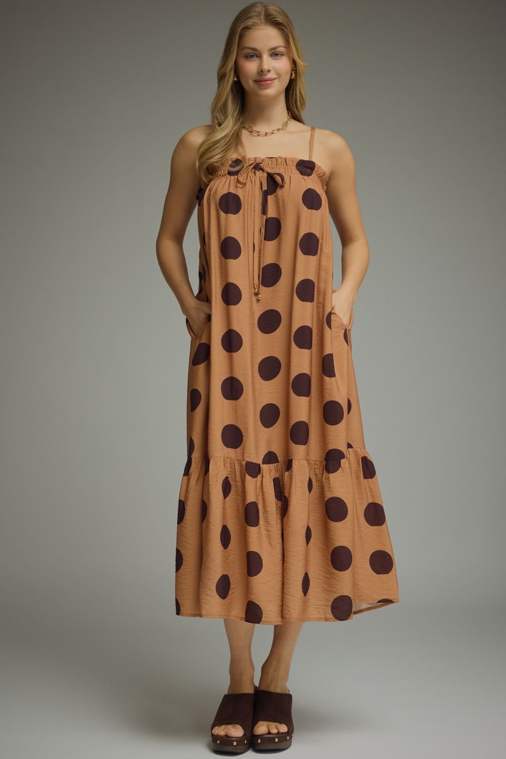 A woman modeling a sleeveless tiered midi dress with a tan base and dark brown polka dots, paired with dark brown slide sandals, against a neutral gray studio background.