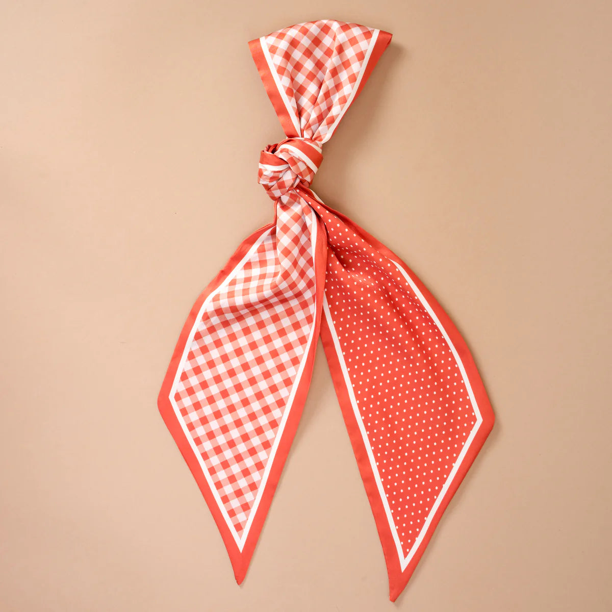 A orange and white silk scarf tied in a knot, featuring a gingham check pattern on one side, a solid red border, and white polka dots on the other, displayed against a solid beige background.