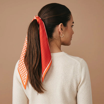 A woman shown from the back wearing a bright orange and white patterned scarf tied around her ponytail, against a solid beige background.