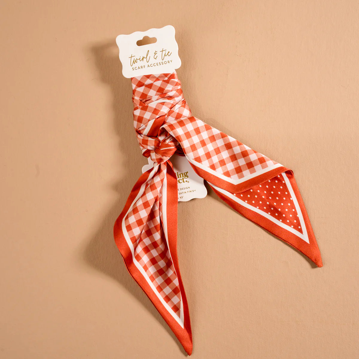 An orange and white checkered neck scarf with solid orange trim, tied in a knot and attached to a white paper hanger tag, displayed on a solid beige background.