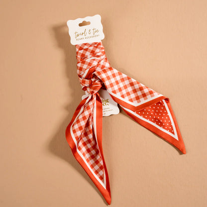 An orange and white checkered neck scarf with solid orange trim, tied in a knot and attached to a white paper hanger tag, displayed on a solid beige background.