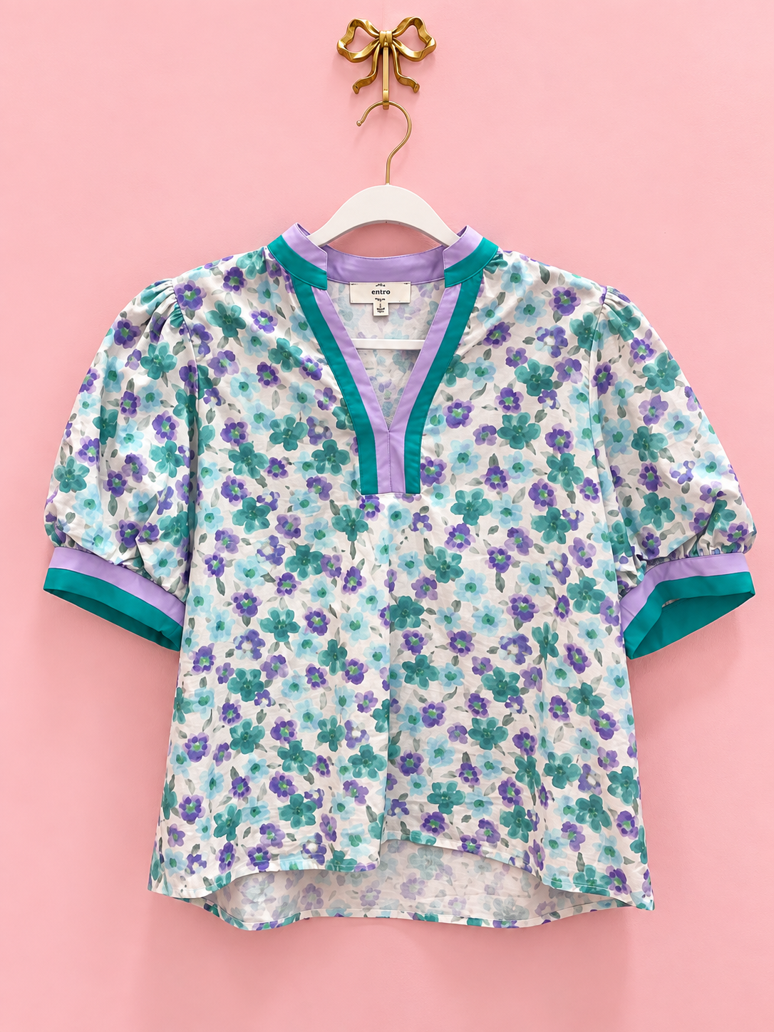 A short-sleeve floral blouse hanging on a white hanger against a solid pink background. The blouse features a white base with purple and teal watercolor-style floral patterns, with solid purple trim along the split neckline and teal trim along the sleeve cuffs.