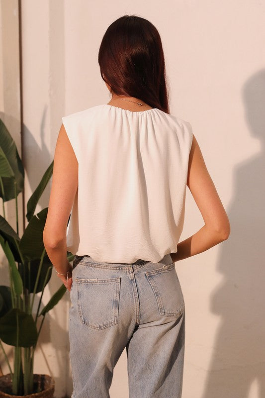 A woman viewed from behind wearing a sleeveless cream-colored top and light-wash blue jeans, standing against a warm-toned wall with a potted plant on the left.