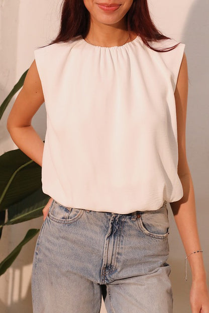 A woman wearing a sleeveless, off-white top with a gathered neckline and light-wash blue jeans, standing against a light-colored wall with a green plant leaf visible on the left.