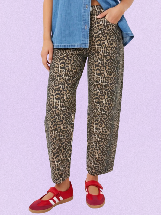 A person wearing leopard-print pants with a blue denim shirt and red shoes, shown from the waist down against a light lavender background.