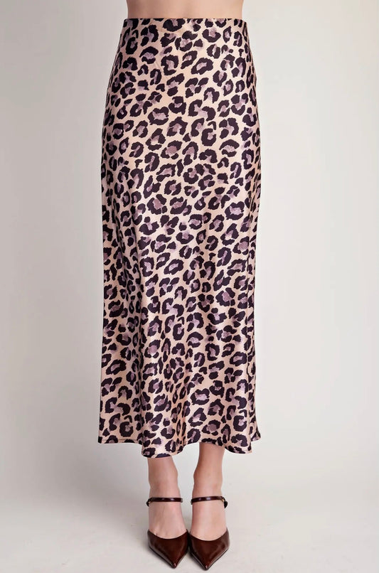A leopard print midi skirt in beige, brown, and black, worn by a model with dark brown pointed-toe heels against a plain light gray studio background.