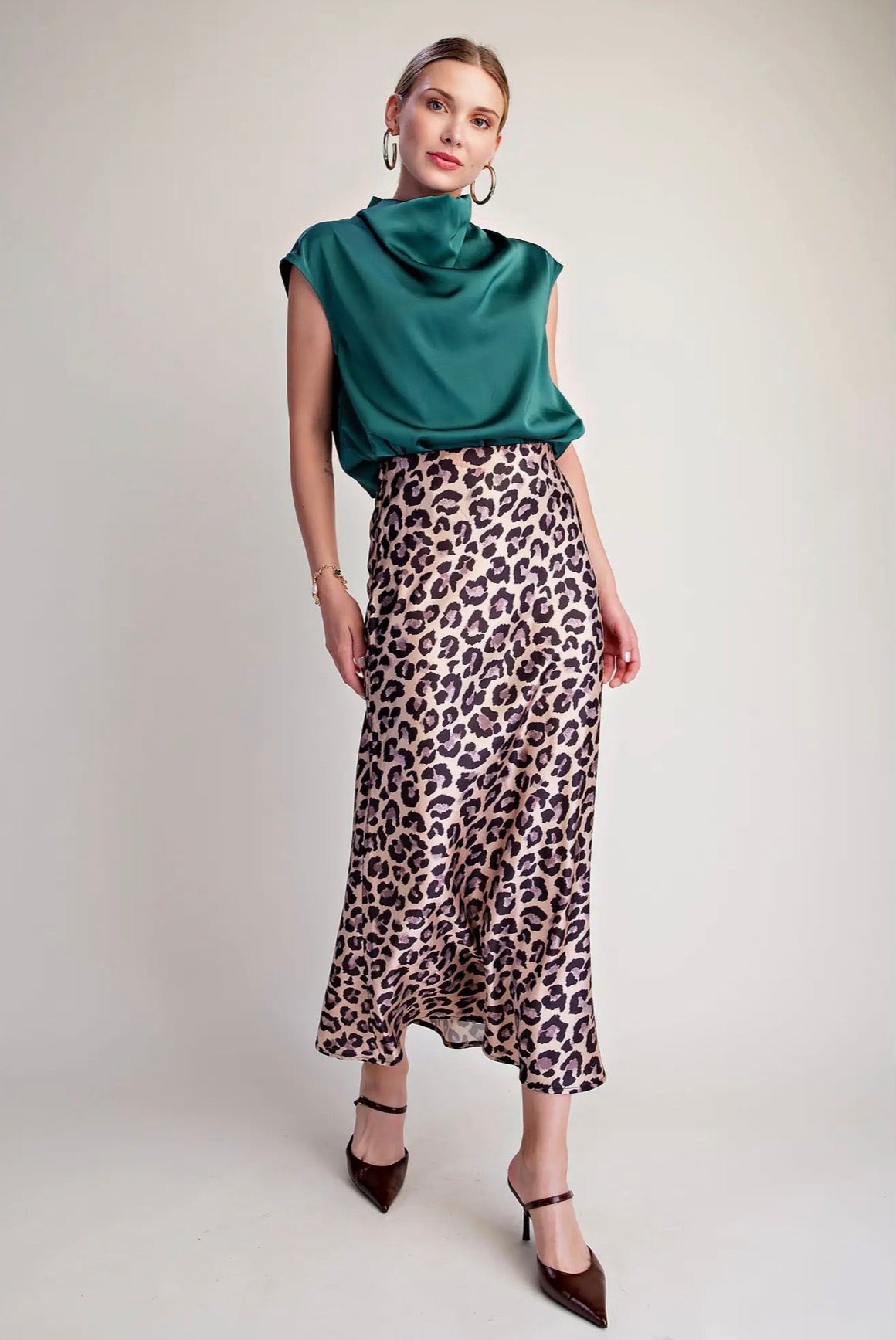 A woman modeling a sleeveless cowl-neck satin top in emerald green paired with a leopard-print midi skirt, brown pointed-toe heels, and large hoop earrings, standing against a plain light gray studio background.