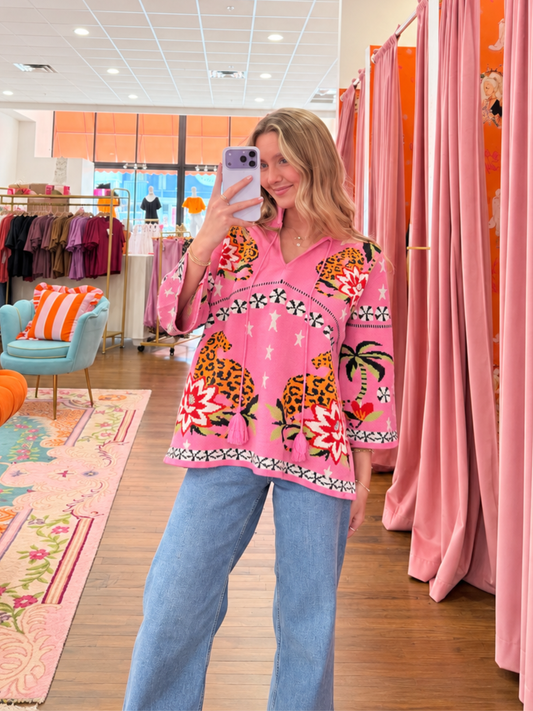 A woman takes a mirror selfie in a clothing boutique wearing a vibrant pink short-sleeve button-up shirt with colorful leopard and floral prints, paired with light blue jeans.