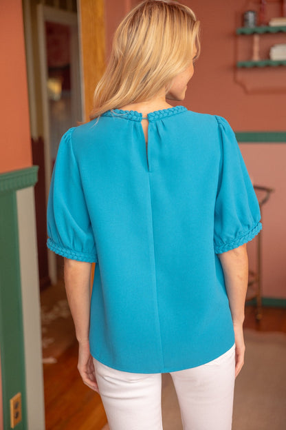Aqua blue short sleeve top with braided trim detail on the neckline and cuffs, featuring a textured fabric and puff sleeves. Stylish and feminine, perfect for casual chic or dressy outfits. Modeled with white jeans in a modern boutique setting.