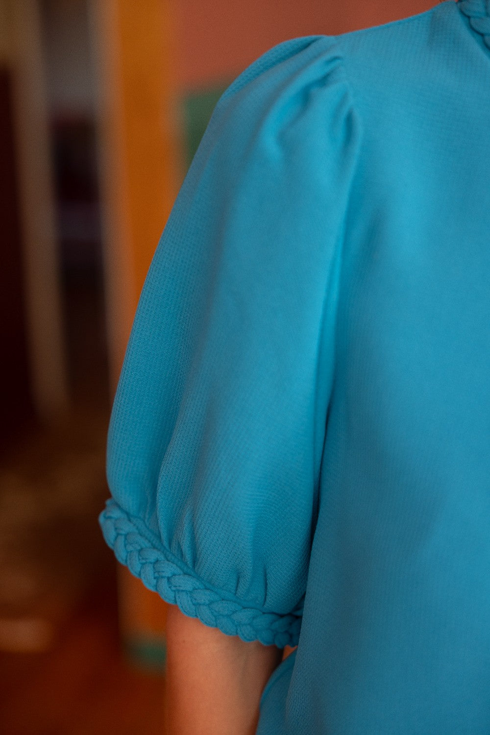 Aqua blue short sleeve top with braided trim detail on the neckline and cuffs, featuring a textured fabric and puff sleeves. Stylish and feminine, perfect for casual chic or dressy outfits. Modeled with white jeans in a modern boutique setting.