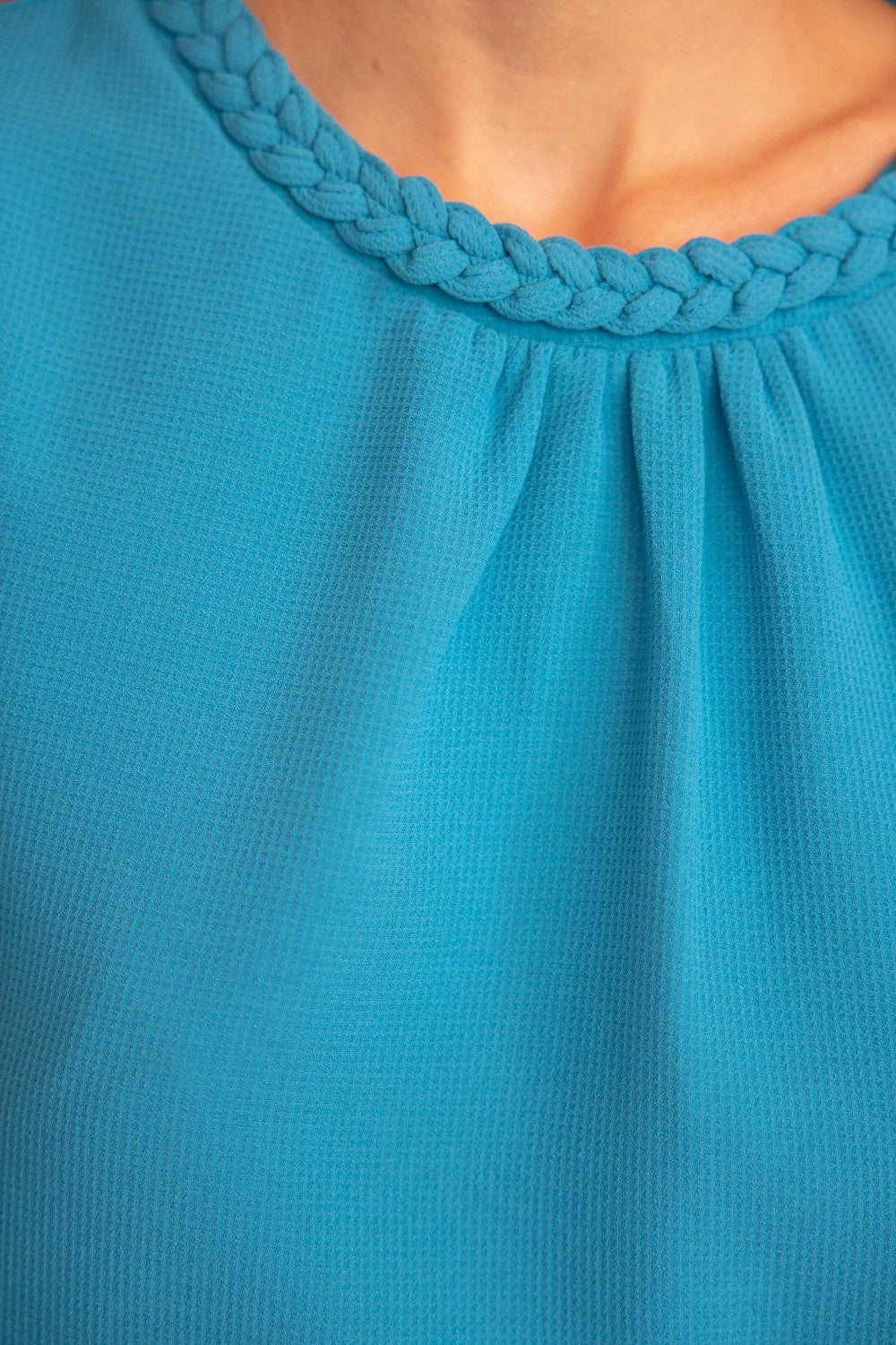 Aqua blue short sleeve top with braided trim detail on the neckline and cuffs, featuring a textured fabric and puff sleeves. Stylish and feminine, perfect for casual chic or dressy outfits. Modeled with white jeans in a modern boutique setting.