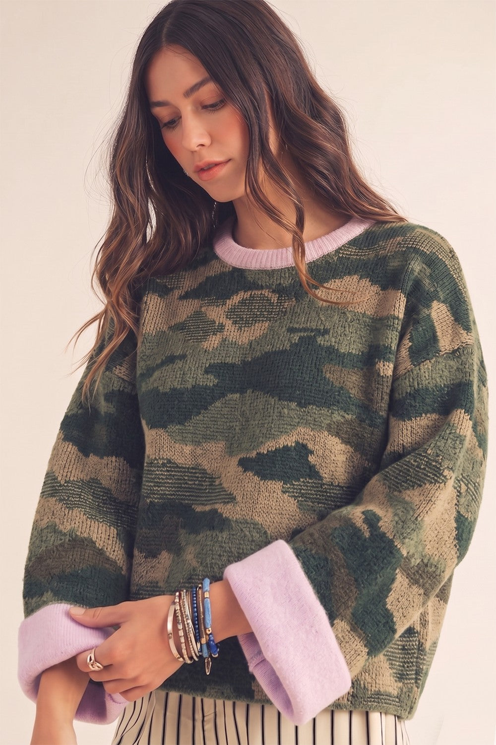 A woman wearing a fuzzy camouflage knit sweater with light purple/lavender ribbed collar and cuffs, paired with striped pants, against a plain light beige background.