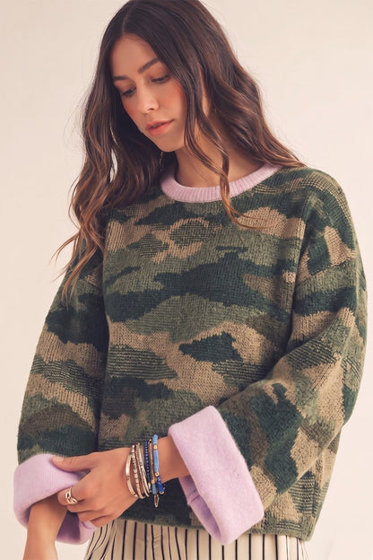 A woman wearing a fuzzy camouflage knit sweater with light purple/lavender ribbed collar and cuffs, paired with striped pants, against a plain light beige background.