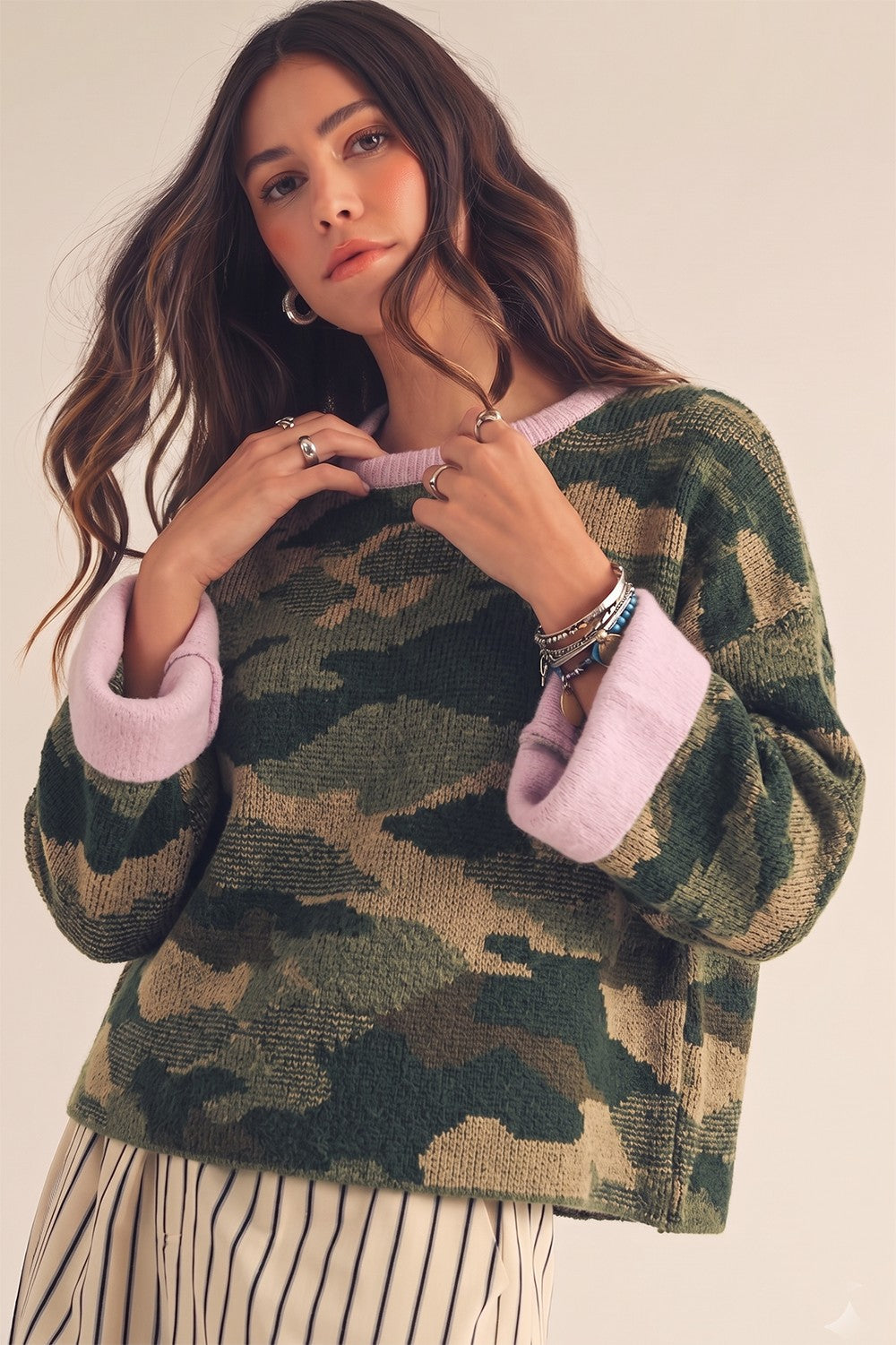 A woman wearing a textured camouflage knit sweater with cream-colored ribbed cuffs and collar, paired with cream and black striped pants, against a plain beige studio background.