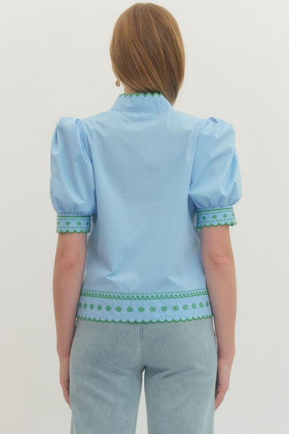 Light blue blouse with green trim worn by a person on a plain background