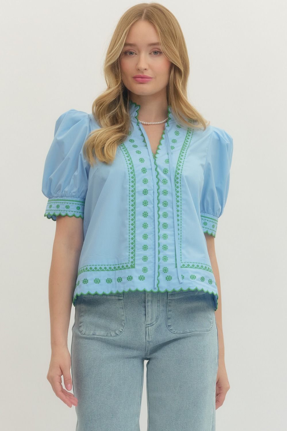 Woman wearing a light blue blouse with green embroidery and jeans on a white background