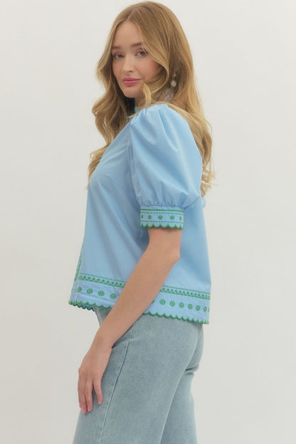 Woman wearing a light blue blouse with green embroidery on a plain background