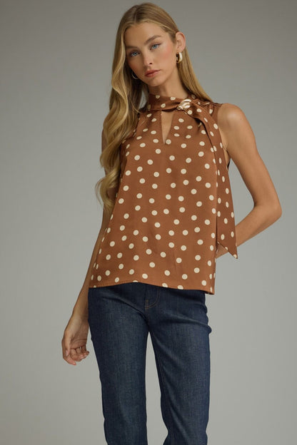 A woman modeling a sleeveless brown top with white polka dots and a tie-neck detail, paired with dark blue jeans, against a solid gray studio background.