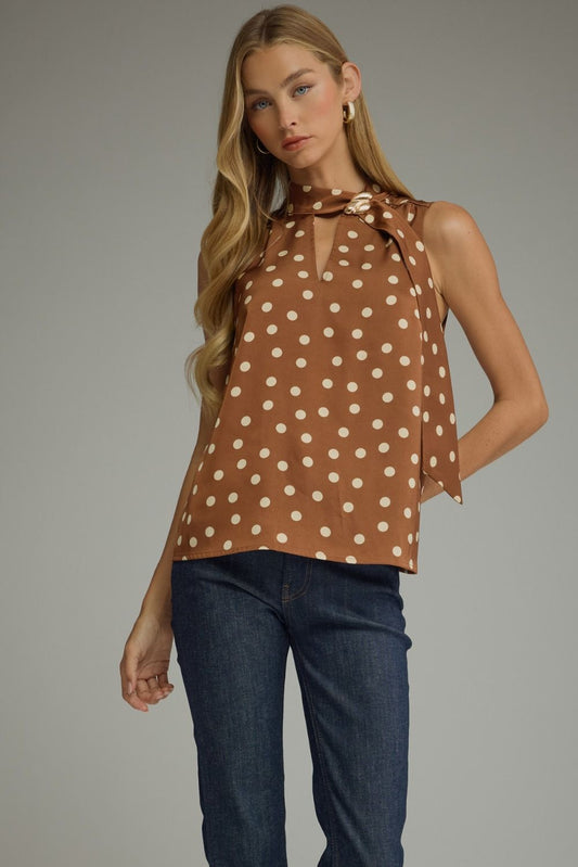 A woman modeling a sleeveless brown top with white polka dots and a tie-neck detail, paired with dark blue jeans, against a solid gray studio background.