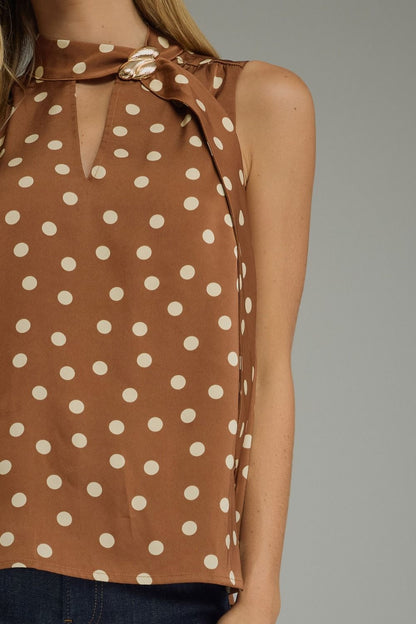 A close-up of a woman wearing a sleeveless brown top with a cream polka dot pattern and a tie-neck detail, paired with dark blue jeans against a solid gray background.