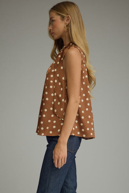 A woman shown in side profile wearing a sleeveless brown top with a white polka dot pattern and blue jeans, photographed against a plain gray studio background.