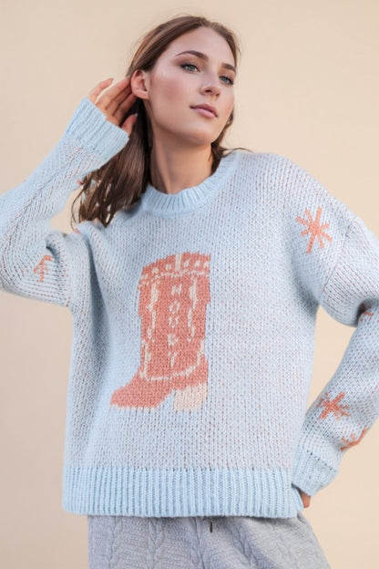 Model wearing The Cozy Cowgirl Sweater — a light blue knit sweater with peach cowgirl boot graphic and star details on the sleeves.