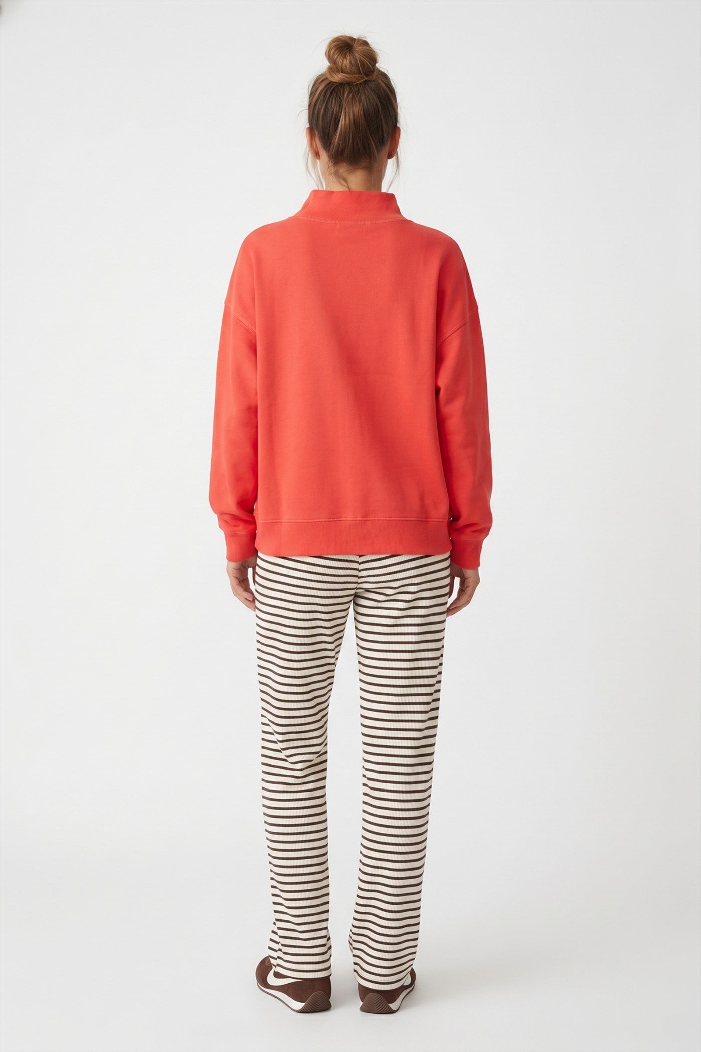 A woman viewed from behind wearing a orange sweatshirt and brown and white striped pants, standing against a light gray studio background.