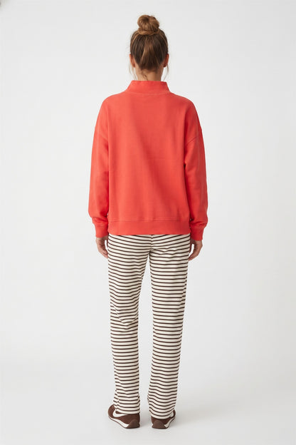A woman viewed from behind wearing a orange sweatshirt and brown and white striped pants, standing against a light gray studio background.