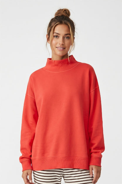 A woman wearing a bright orange mock neck sweatshirt and black and white striped pants, standing against a light gray studio background.