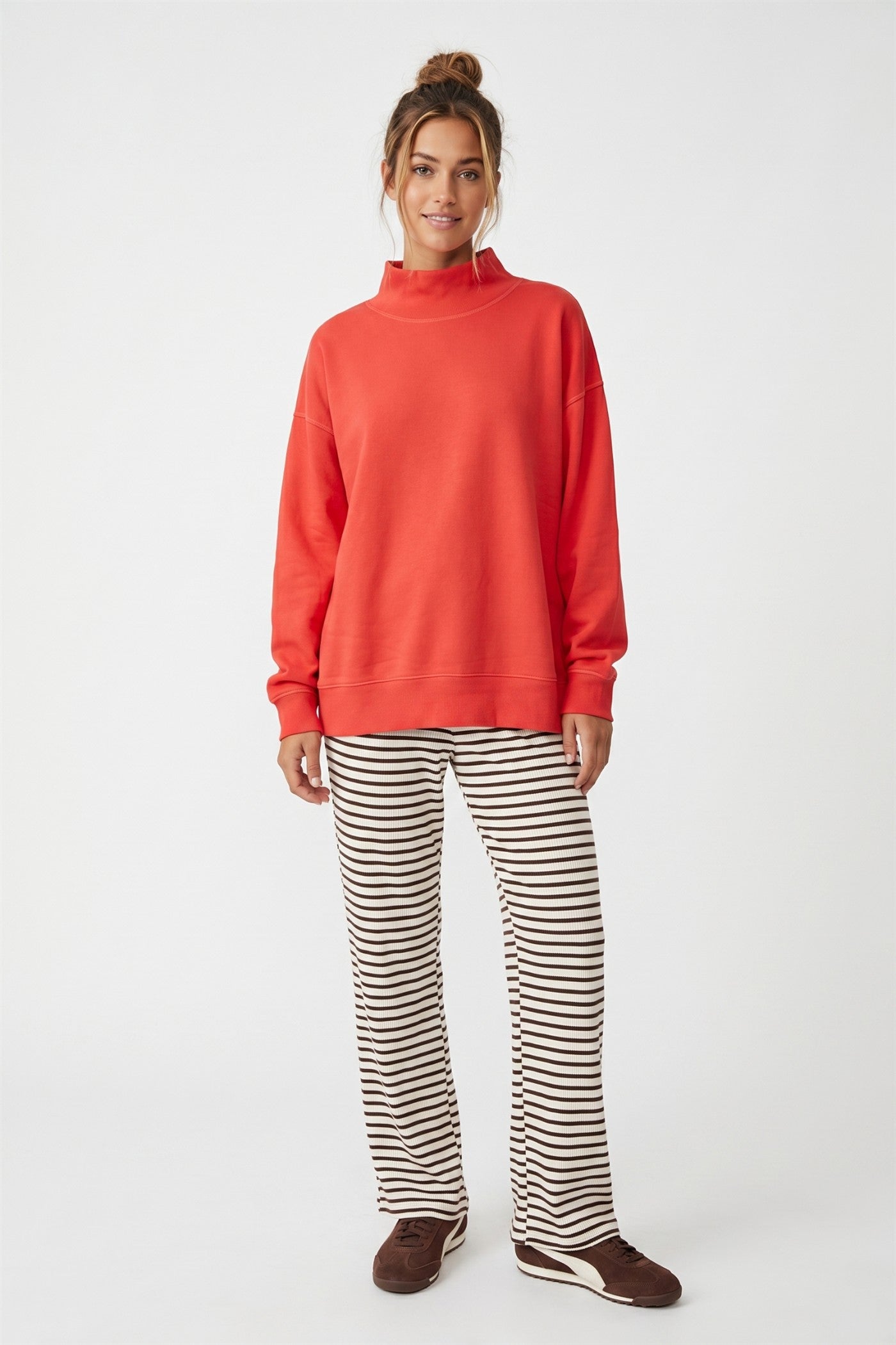 A woman modeling a bright orange mock-neck sweatshirt and brown-and-white horizontally striped pants against a plain white studio background.