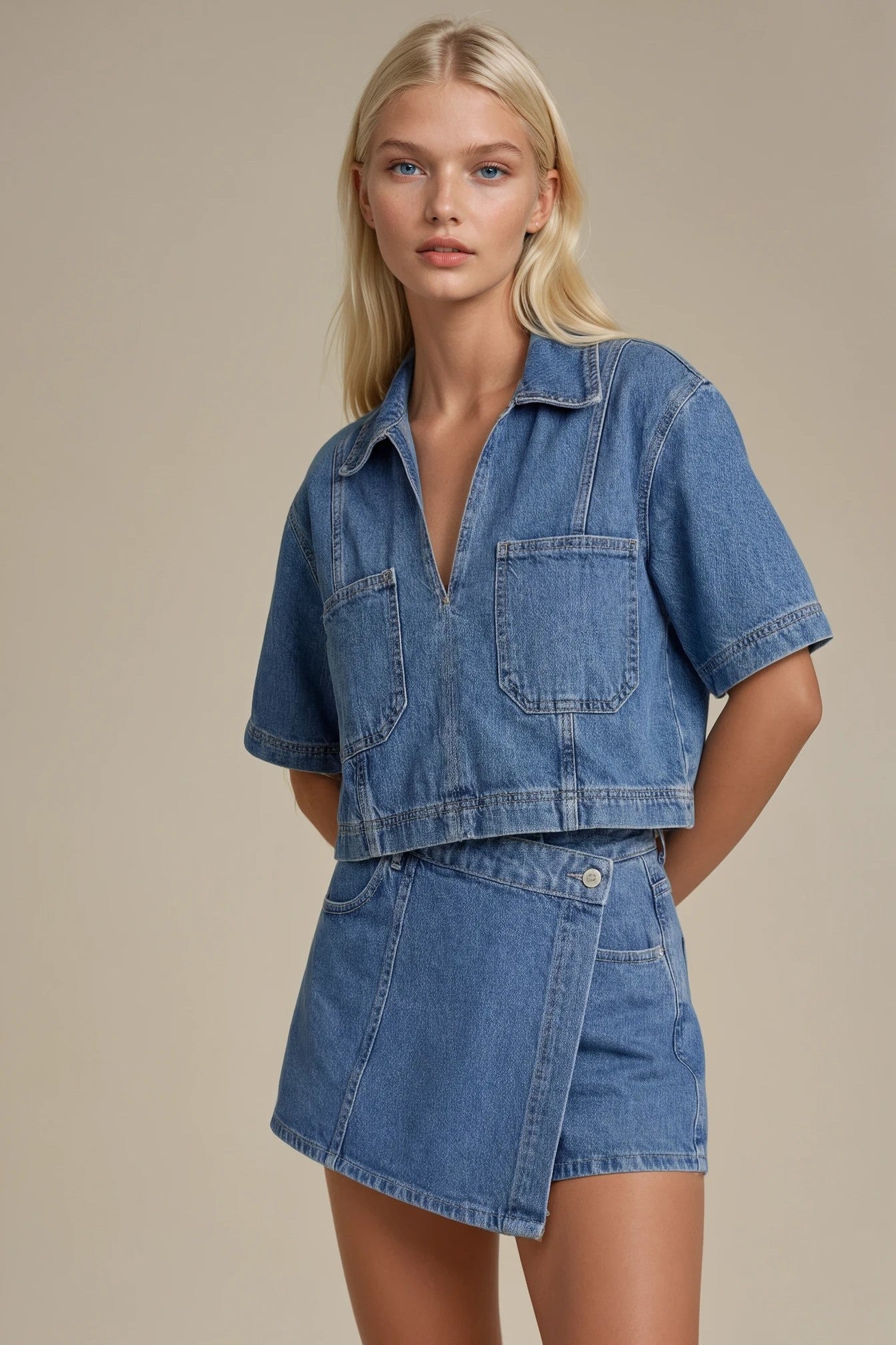 A blonde model wearing a matching blue denim set consisting of a short-sleeve cropped button-front shirt and an asymmetrical wrap mini skirt, photographed against a solid beige studio background.