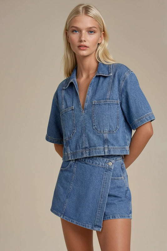 A blonde model wearing a matching blue denim set consisting of a short-sleeve cropped button-front shirt and an asymmetrical wrap mini skirt, photographed against a solid beige studio background.