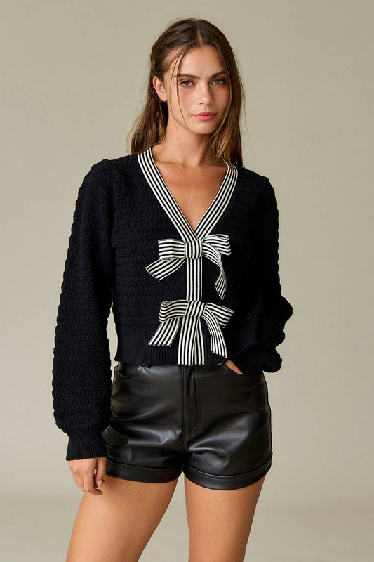 Black textured knit sweater with two black-and-white striped statement bows on the front and a deep V-neckline, styled with black leather shorts