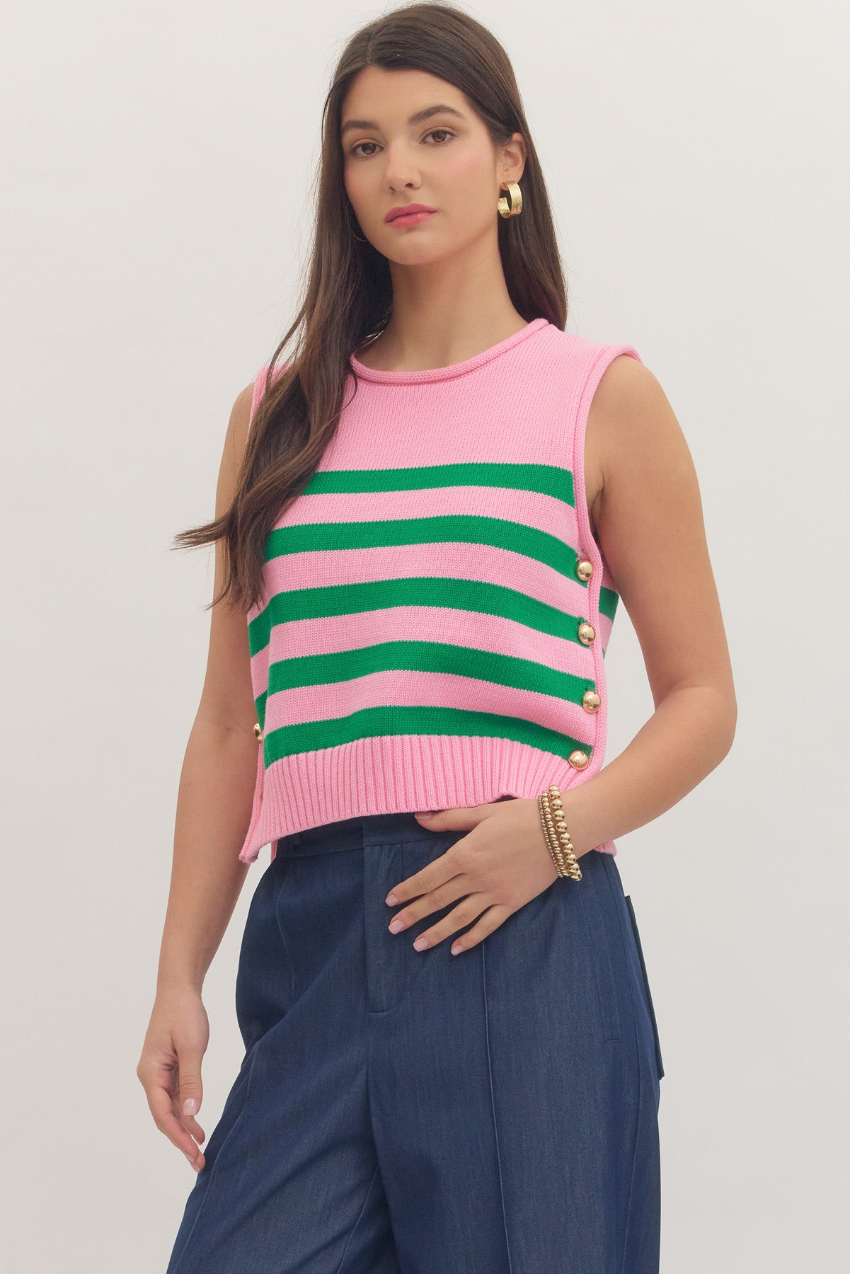 Woman wearing a pink and green striped sleeveless top with blue pants on a plain background