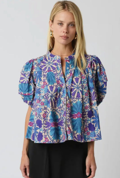 Model wearing a puff sleeve floral blouse in shades of cobalt blue, violet, and berry on a cream background, featuring a button neckline and flowy fit from Milo & Lily Boutique