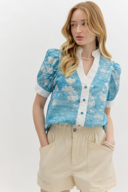 A woman with wavy blonde hair wears a blue floral watercolor puff sleeve blouse with a white mandarin collar and button-front detail. The blouse features a soft, airy fabric with a Monet-inspired water lily print. She pairs it with beige high-waisted shorts and pearl accessories, posing against a clean white background.